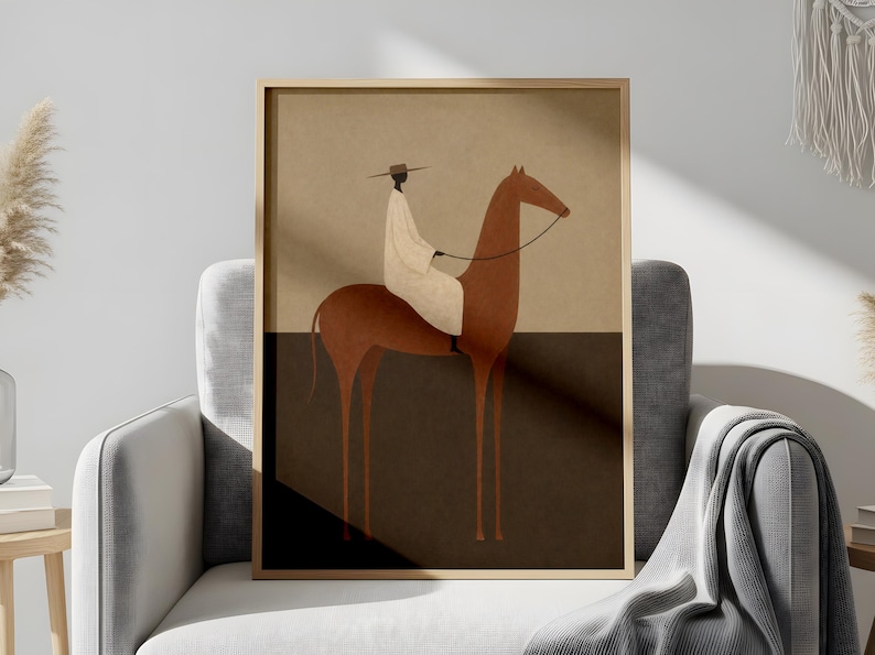 May include: Framed art print featuring an illustration of a person riding a horse. The rider is wearing a white garment and hat. The horse is brown, and the background is divided into tan and dark brown sections. The artwork is displayed in a gold frame.