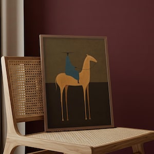 May include: A framed art print featuring a minimalist illustration of a camel with a rider. The artwork uses a palette of muted earth tones, with a blue-clad figure atop the camel. The print is displayed on a wooden chair with a woven seat.