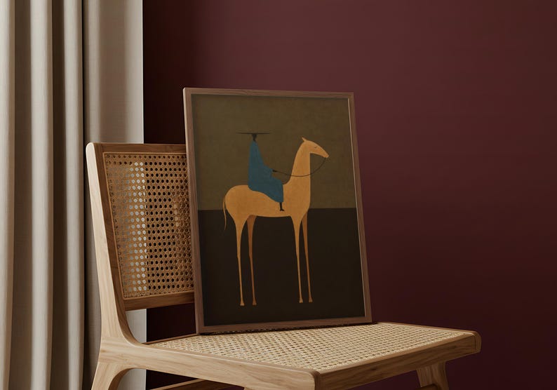 May include: A framed art print featuring a minimalist illustration of a camel with a rider. The artwork uses a palette of muted earth tones, with a blue-clad figure atop the camel. The print is displayed on a wooden chair with a woven seat.