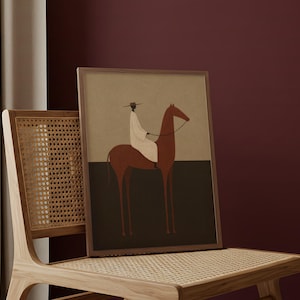 May include: A framed art print featuring a minimalist illustration of a rider on a horse. The artwork uses a muted color palette with brown, black, and beige tones. The print is displayed on a wooden chair with a woven seat and back.