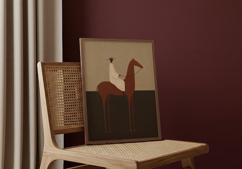 May include: A framed art print featuring a minimalist illustration of a rider on a horse. The artwork uses a muted color palette with brown, black, and beige tones. The print is displayed on a wooden chair with a woven seat and back.
