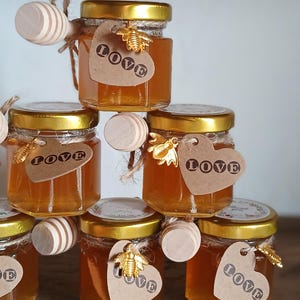 May include: A pyramid of small glass jars filled with golden honey. Each jar has a gold lid, a heart-shaped tag with the word "love", and a wooden honey dipper. A gold bee charm is attached to some of the tags.