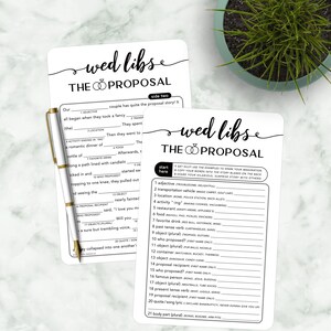 Könnte beinhalten: Zwei weiße "Wed Libs: The Proposal"-Karten mit schwarzem Text und einem goldenen Stift. Die Karten enthalten Ausfüllaufforderungen für eine Heiratsantragsgeschichte. Die obere Karte hat eine teilweise ausgefüllte Geschichte. Die untere Karte hat eine Liste mit Wortaufforderungen.
