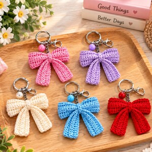 Può includere: Cinque portachiavi a forma di fiocco all'uncinetto nei colori rosa, viola, crema, blu e rosso. Ogni portachiavi è dotato di un anello in metallo, un moschettone e una perlina rotonda abbinata alla catenella. I fiocchi hanno una trama a maglia visibile.