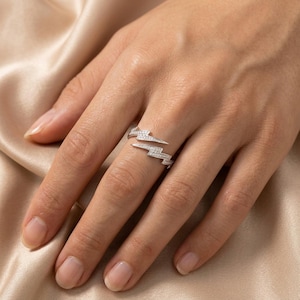 May include: A silver-colored ring in a lightning bolt design, encrusted with small, clear gemstones. The ring is worn on a finger, showcasing its wrap-around style and sparkling details. The ring is set against a neutral background.