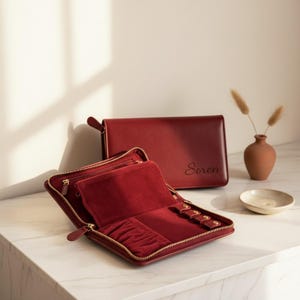 Pode incluir: Um estojo organizador de joias retangular vermelho escuro com fecho de correr. O exterior é liso com o nome Soren gravado na frente. O interior é forrado com veludo vermelho a condizer e possui vários compartimentos e abas com botões.