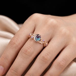 Può includere: Anello in oro rosa con una grande gemma rotonda color teal incastonata in un design a mezzaluna. La fascia è adornata da piccole gemme viola e dettagli a forma di foglia, creando un'estetica elegante e ispirata alla natura.