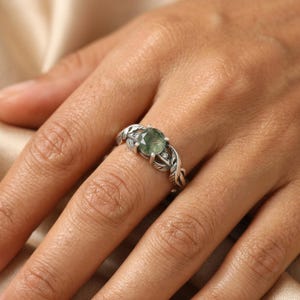 May include: A silver ring with a round, green gemstone. The ring has a nature-inspired design with leaf and vine detailing. Small, clear accent stones are set around the main stone. The ring is worn on a finger.