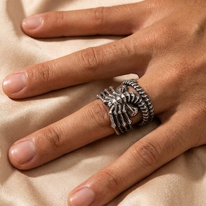 May include: A silver-toned ring featuring a detailed spider design with elongated legs that wrap around the finger. The metal has an oxidized finish to highlight the textured segments of the spider's body and legs, creating a distinct contrast.