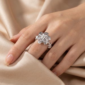 以下が含まれることがあります： 銀色のリングで、花のモチーフが特徴です。リングの焦点は、繊細な花びらと小さな輝くアクセントを持つ多層の花です。バンドは自然にインスパイアされたデザインです。