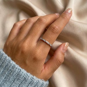 Op de afbeelding: Een zilveren ring met een gedraaid ontwerp, bezet met kleine, heldere edelstenen. De ring wordt aan een vinger gedragen, met een lichtblauwe mouw in beeld. De achtergrond is een zachte, neutrale kleur.