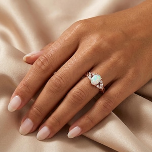 May include: Rose gold ring featuring an oval opal gemstone. The ring has a delicate, swirling design with small, clear accent stones. The opal has a milky white appearance with subtle color variations. The ring is displayed on a hand with manicured nails.
