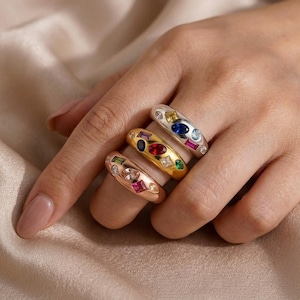 May include: Three rings with colourful gemstones. One is silver with blue, yellow, and clear stones. The second is gold with red, blue, and clear stones. The third is rose gold with pink, green, and clear stones.
