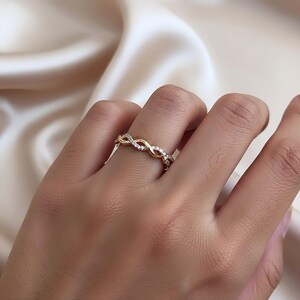 Könnte beinhalten: Ein goldener Ring mit einem gedrehten Unendlichkeitsdesign. Ein Strang des Rings ist aus poliertem Gold, während der andere Strang mit kleinen, klaren, funkelnden Steinen besetzt ist, die dem durchgehenden, verschlungenen Muster folgen.