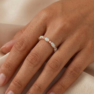 May include: A rose gold band ring featuring a series of alternating oval-shaped opals and small round clear stones. The opals display iridescent flashes of colour, and the metal band has a polished finish, creating a delicate and refined appearance.