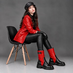 Könnte beinhalten: Ein roter Blazer mit Krokodilprägung, eine schwarze Hose in Lederoptik und schwarze Handschuhe. Das Outfit umfasst eine schwarze Schiebermütze und wadenhohe Stiefel mit rotem Obermaterial in Krokodiloptik und einer schwarzen Profilsohle.