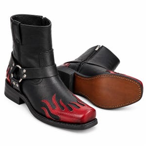 Puede incluir: Botines de cuero negro con punta cuadrada y detalles de llamas rojas. Las botas incluyen una correa de arnés con un anillo de metal plateado y remaches, cierre de cremallera lateral y una suela marrón con costuras visibles en el borde.