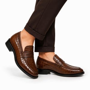 Puede incluir: Un par de mocasines marrones con un patrón de relieve de cocodrilo, una tira clásica sobre el empeine y un tacón y suela de color negro. Los zapatos se llevan con pantalones de color marrón oscuro que terminan por encima del tobillo.