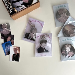 May include: A selection of anime-style merchandise, including pin badges, acrylic keychains, and small art prints. The items feature illustrations of male characters. A box with the text "Codename Anastasia" is also present.