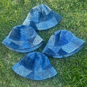 May include: Four blue patchwork bucket hats are displayed on green grass. Each hat features a different arrangement of patterned fabric sections. The hats have a classic bucket hat shape with a slightly sloped brim. The fabric appears to be denim or a similar material.