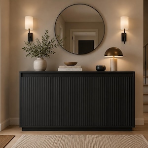 May include: A black rectangular console table with a vertical ribbed texture on the front doors. The top surface holds a light-colored vase with green foliage, a small wooden bowl, two stacked books, a small black bowl, and a dome-shaped lamp.