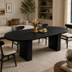 May include: An oval black wooden dining table with a fluted pedestal base, accompanied by four black chairs featuring light beige upholstered seats. A yellow vase with dried flowers and a small plate of bread sit on the table surface.