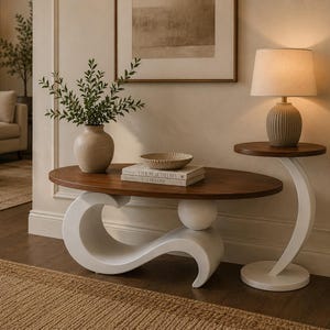 May include: An oval wooden coffee table with a white, sculptural, wave-shaped base and a matching white side table with a circular wooden top. The coffee table holds a beige vase with green foliage, a shallow bowl, and two stacked books.