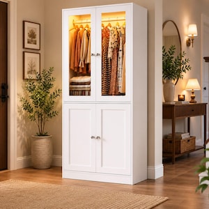 May include: A tall white storage cabinet featuring two upper glass-paneled doors with internal lighting and two lower solid doors. The upper section contains a hanging rod with various garments and folded items, while the lower section provides enclosed storage.