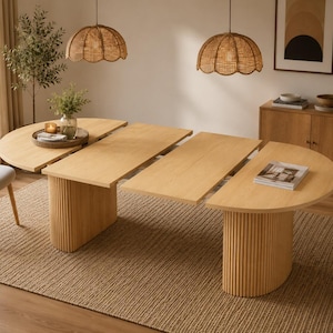 May include: An extendable light wood dining table featuring a smooth rectangular top with two semi-circular ends and two central rectangular leaves. The table is supported by two cylindrical bases with vertical fluted detailing.