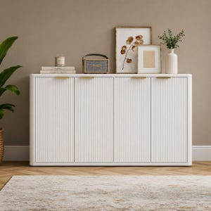 May include: A white sideboard featuring four doors with a vertical fluted texture and thin gold horizontal handles. The top surface holds a small speaker, two stacked books, a candle, two framed botanical prints, and a white vase with greenery.