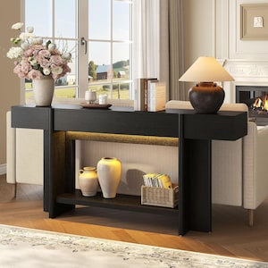May include: A long, black wooden console table with two drawers and an open lower shelf. The table features integrated warm under-shelf lighting. It is decorated with a vase of flowers, books, a table lamp, two ceramic vases, and a woven basket.