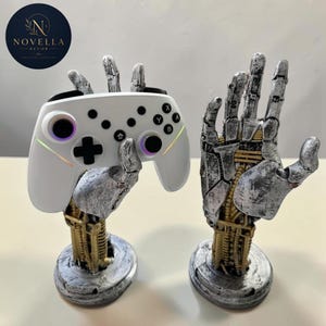 Könnte beinhalten: Zwei roboterartige Hand-Gaming-Controller-Ständer. Einer hält einen weißen Controller mit schwarzen Knöpfen und leuchtenden Akzenten. Der andere Ständer ist leer. Die Ständer sind silber- und goldfarben, mit rundem Sockel. Das Novella Decor-Logo befindet sich oben links.