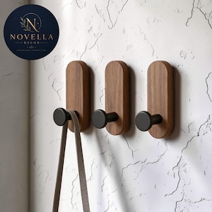 Puede incluir: Tres ganchos de pared con placas traseras de madera ovaladas y verticales con veta natural y pernos cilíndricos de color negro mate. Una correa fina de color marrón claro cuelga de un gancho. El logo circular dice NOVELLA DECOR.