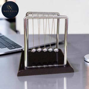 May include: A Newton's cradle desk toy with five polished steel balls suspended from a chrome frame. The base is dark brown, and the logo "Novella Decor" is visible in the upper left corner. This executive desk accessory is designed for stress relief.