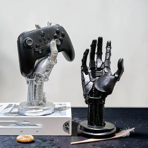 Könnte beinhalten: Zwei Roboterhand-Skulpturen. Eine silberne Hand hält einen schwarzen Videospiel-Controller. Die andere ist eine schwarze Roboterhand. Beide stehen auf kreisförmigen Sockeln. Die silberne Hand steht auf einem weißen Buch.