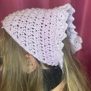 May include: A light purple crochet bonnet with a scalloped edge. The bonnet is made of a soft, open-weave material and is worn on the head. The bonnet is a light purple color.