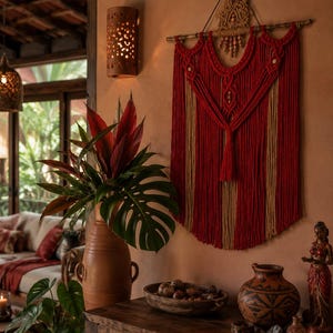 May include: A wall hanging made of red and tan macrame cord suspended from a wooden rod. The design features intricate knotted patterns, hanging fringe, and a central tassel. The piece is displayed against a warm-toned wall, showcasing a layered fiber art construction with decorative wooden beads integrated into the upper section.