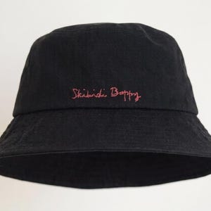 Op de afbeelding: Zwarte vissershoed met een brede rand. De hoed heeft de geborduurde tekst "Skibidi Boppy" in roze script over de voorkant. De hoed is gemaakt van een zachte, donkere stof.