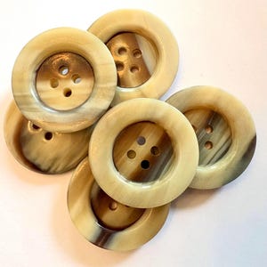 May include: A collection of six round, four-hole buttons featuring a smooth, polished surface with a marbled pattern in shades of cream, beige, and dark brown. Each button has a wide, raised outer rim and a recessed center area.