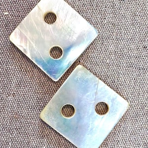 May include: Two square-shaped buttons crafted from iridescent mother-of-pearl material. Each button features a smooth, reflective surface with natural pearlescent color variations and two circular holes positioned vertically for attachment purposes.