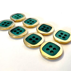 Op de afbeelding: Een set van acht ronde knopen, elk voorzien van een gepolijste goudkleurige metalen rand rondom een vierkant blauwgroen inlegstuk. Elke knoop heeft vier kleine gaatjes die in een vierkant patroon in het midden van het oppervlak zijn geplaatst.