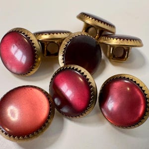 May include: A collection of round buttons featuring smooth, deep red, translucent centres set within textured, brass-toned metal rims. Each button has a shank on the back for attachment, with the red surfaces displaying a subtle, glossy sheen.