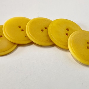 May include: Five round, bright yellow buttons arranged in a slight arc. Each button features a smooth, raised centre section surrounded by a flat outer rim and contains two small, vertically aligned holes in the middle for attachment purposes.