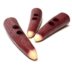May include: Three elongated toggle buttons with a textured, dark reddish-brown surface and a smooth, light cream-coloured tip. Each button features two circular holes positioned along the centre for attachment to a garment or accessory.