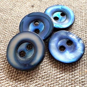 Op de afbeelding: Vier ronde knopen met een glanzende, iriserende blauwe afwerking. Elke knoop heeft een glad, licht concaaf oppervlak en twee centrale gaten voor bevestiging. De knopen vertonen verschillende tinten diep marineblauw en helder kobaltblauw.
