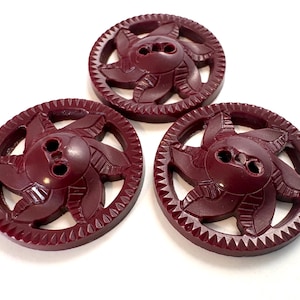 May include: Three circular burgundy buttons featuring a central two-hole design surrounded by a stylized, radiating petal pattern. Each button has a serrated, gear-like outer edge and a smooth, glossy finish, arranged in a triangular formation.