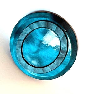 May include: A circular button with a glossy, iridescent teal finish. The design features a smooth, raised central disc surrounded by a textured ring with subtle, radiating geometric facets, creating a layered appearance with deep blue and green tones.