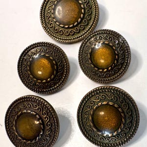 May include: Five round buttons featuring a central amber-coloured dome surrounded by a metallic bronze-toned border. The border displays a raised rope-like edge and an intricate swirling scroll pattern, creating a textured and layered appearance.