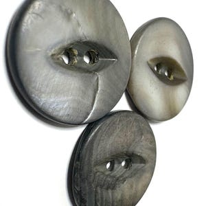 May include: Three circular buttons with a smooth, iridescent grey finish. Each button features a recessed, almond-shaped centre containing two holes for attachment. The buttons vary slightly in size and display natural, swirling patterns.