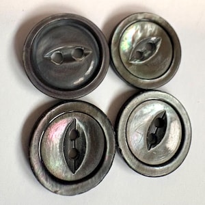 Pode incluir: Quatro botões circulares com um acabamento cinza escuro iridescente. Cada botão apresenta um centro rebaixado com dois orifícios dispostos verticalmente dentro de um recorte alongado em forma de olho. A superfície exibe um brilho sutil.